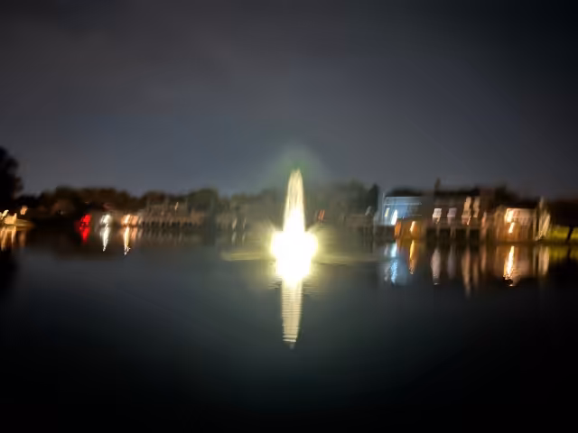 Blurry image of a small lake at night. There are houses surrounding the lake. In the center of the lake is a lit up fountain in the vague shape of a penis with a larger stream of water in the center and smaller jets on the left and right of the larger stream.